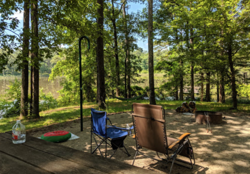A serene campsite by a lake, featuring two chairs, a fire pit, and surrounded by lush green trees.