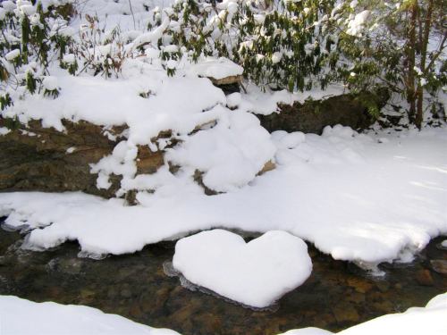 A heart-shaped patch of snow rests on a rocky surface beside a gently flowing stream in a winter landscape.