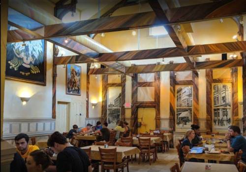 A cozy restaurant interior with wooden beams, murals, and patrons dining at tables. Warm lighting creates a welcoming atmosphere.