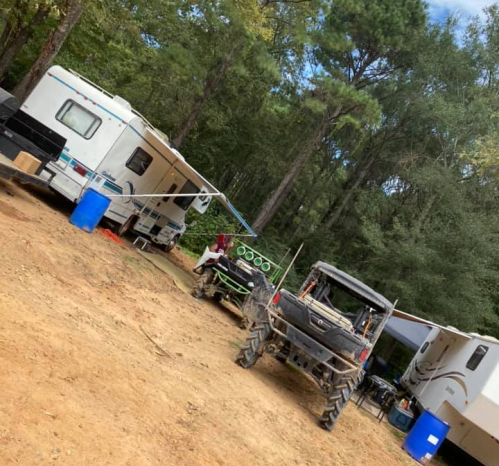 Two RVs parked in a wooded area, with a utility vehicle and outdoor equipment nearby on a dirt path.
