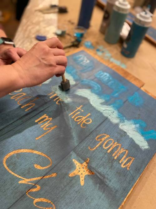 A person paints a wooden sign with blue and gold lettering, featuring waves and a starfish design.