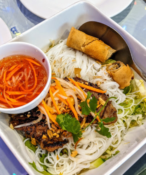A bowl of vermicelli noodles topped with grilled meat, fresh vegetables, herbs, and a side of dipping sauce.