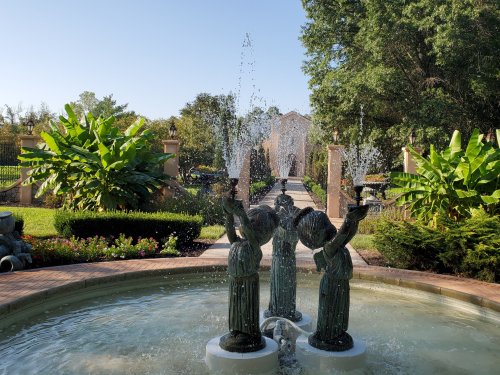 A fountain featuring three cherubic statues, surrounded by lush greenery and a pathway leading to a building in the background.