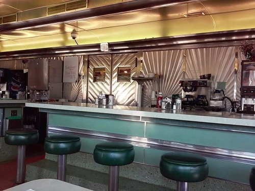 A retro diner interior featuring a green counter, metal accents, and empty stools, evoking a nostalgic atmosphere.