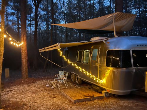A silver Airstream trailer at dusk, adorned with string lights and a canopy, surrounded by trees.