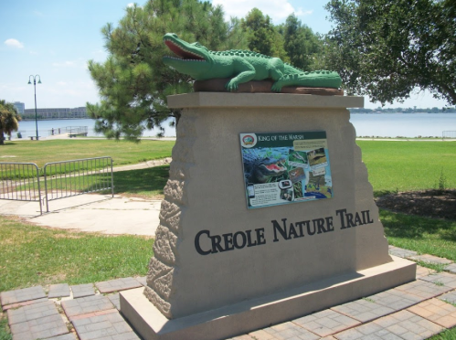 Sign for Creole Nature Trail featuring a crocodile sculpture, with a scenic view of water and greenery in the background.