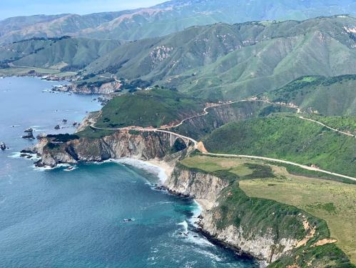 Aerial view of a coastal landscape with green hills, cliffs, and a winding road along the shoreline.