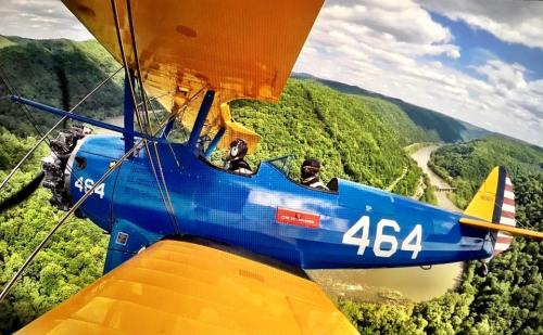 A blue biplane with yellow wings flying over lush green hills and a winding river below.