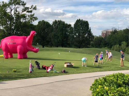 A large pink inflatable elephant stands on a golf course, while children practice putting nearby.