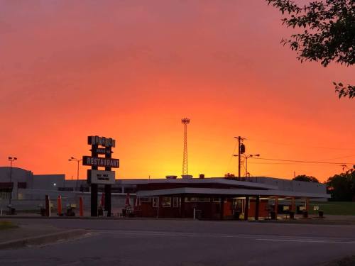 A restaurant sign stands in front of a vibrant orange sunset, with silhouettes of trees and a tower in the background.