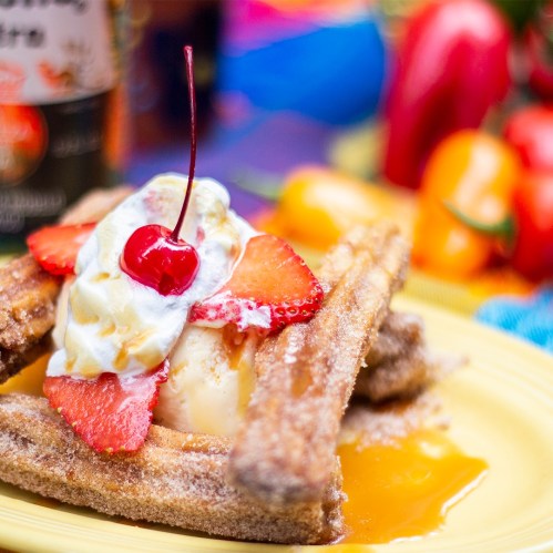 A plate of churros topped with ice cream, strawberries, whipped cream, and a cherry, with colorful background elements.