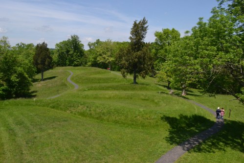 A winding path through lush green hills and trees under a clear blue sky, with people walking in the distance.