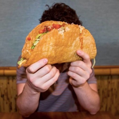 A person holds a large taco in front of their face, with colorful toppings visible inside the taco shell.