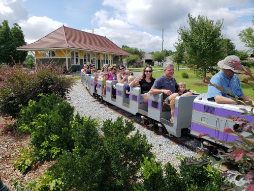 A miniature train with passengers rides along a gravel path near a colorful station surrounded by greenery.
