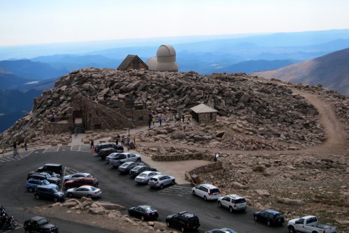 A rocky mountain summit with an observatory, parking area, and visitors enjoying the scenic view.