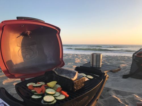 A portable grill on the beach with vegetables cooking, overlooking the ocean at sunset.