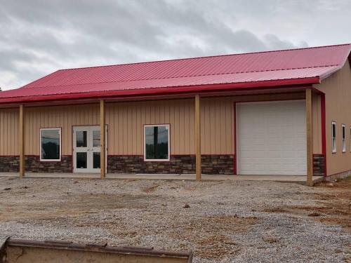 A new building with a red metal roof, stone accents, and large windows, set on a gravel lot under a cloudy sky.