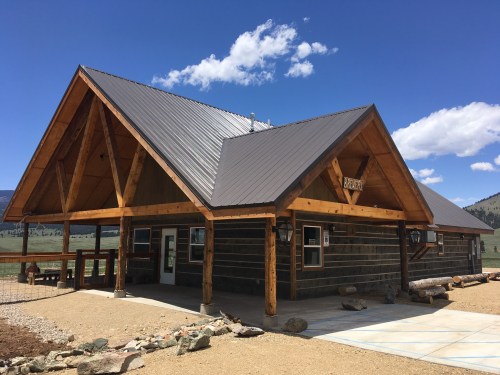 A rustic wooden cabin with a sloped roof, large overhangs, and a stone pathway, set against a blue sky and mountains.