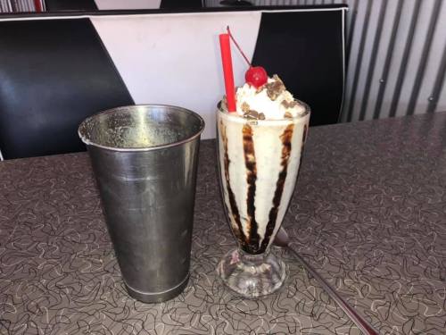 A tall glass of ice cream topped with whipped cream, a cherry, and chocolate syrup, next to a metal milkshake cup.
