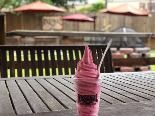 A soft-serve ice cream in a cup on a wooden table, with outdoor seating and umbrellas in the background.