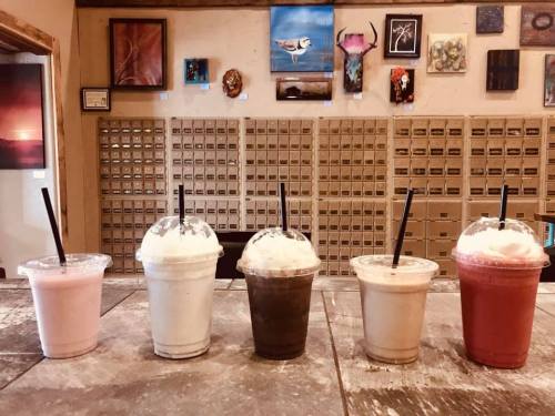 Five colorful milkshakes with whipped cream sit on a counter, against a backdrop of art and storage boxes.
