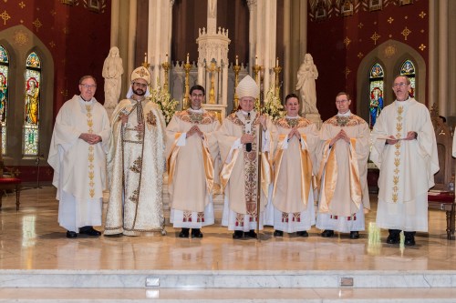 A group of clergy in ceremonial attire stands in a church, with an altar and stained glass windows in the background.