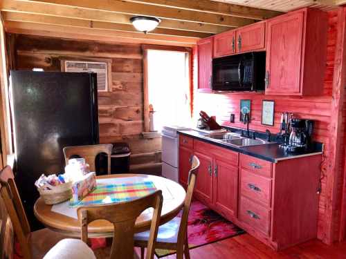 Cozy kitchen with red cabinets, black appliances, a dining table, and wooden walls, illuminated by natural light.