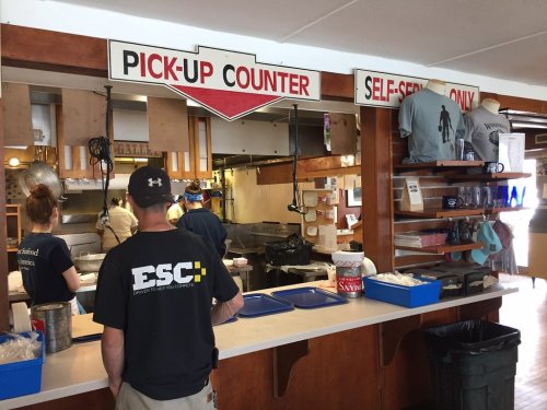 A person stands at a pick-up counter in a casual dining area with signs for self-service and food preparation in the background.