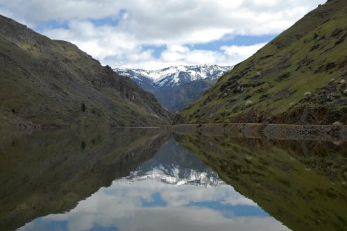 A serene mountain landscape with a calm lake reflecting snow-capped peaks and lush green hills under a cloudy sky.