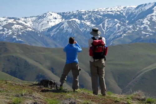 Two hikers stand on a hillside, gazing at snow-capped mountains in the distance under a clear blue sky.