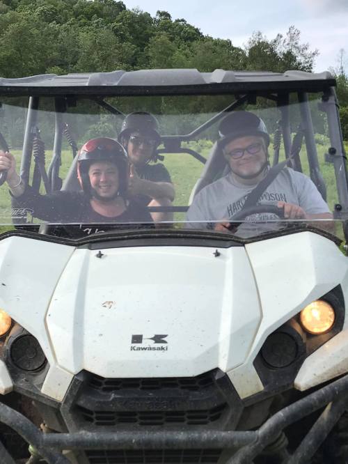 Three people smiling in a Kawasaki off-road vehicle, wearing helmets, with greenery in the background.
