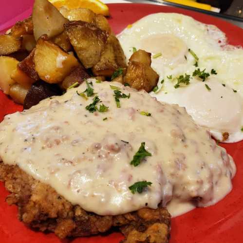 A red plate with fried chicken smothered in gravy, two fried eggs, and seasoned roasted potatoes.