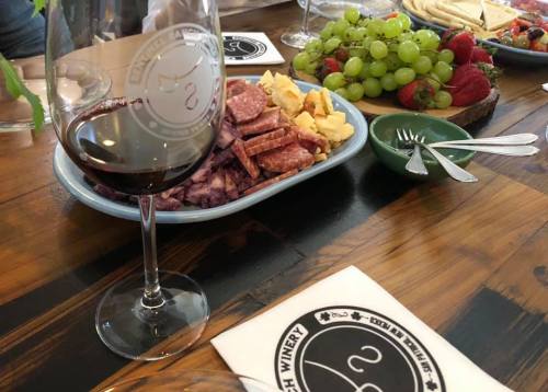 A glass of red wine beside a platter of meats, cheese, and fresh fruit on a wooden table.