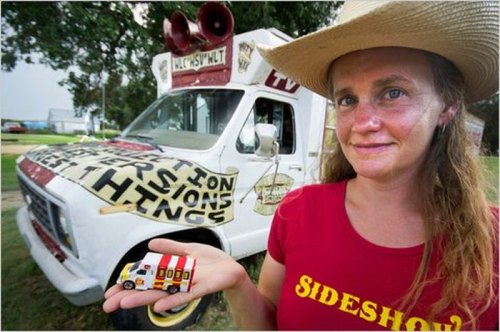 A woman in a cowboy hat holds a toy truck in front of a colorful, sign-covered van promoting a sideshow.