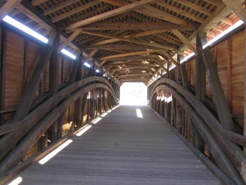 Interior view of a wooden covered bridge, showcasing arched beams and a wooden floor, leading to bright light at the end.