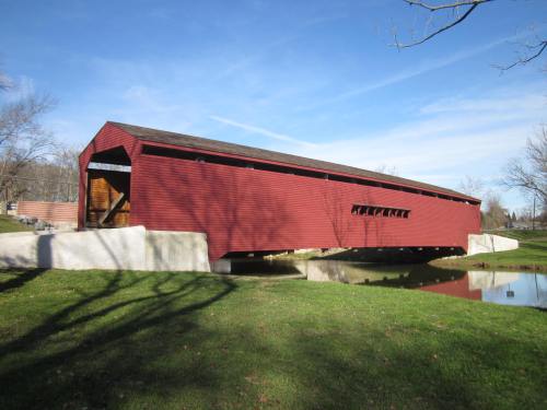 A red covered bridge spans a small stream, surrounded by green grass and bare trees under a clear blue sky.