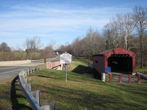 A red covered bridge beside a road, with a historical marker and trees in the background under a clear blue sky.