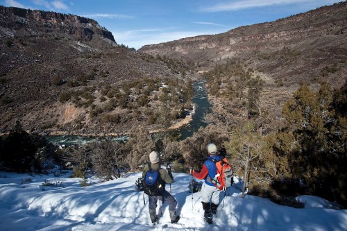 Two hikers in winter gear stand on a snowy ridge, overlooking a winding river and rugged landscape below.