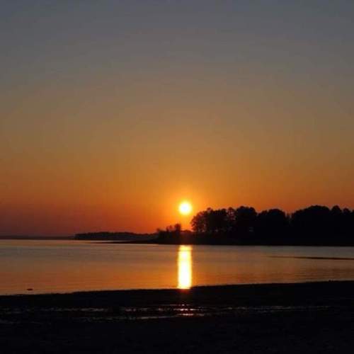 A serene sunset over a calm lake, with silhouettes of trees and a warm orange sky reflecting on the water.