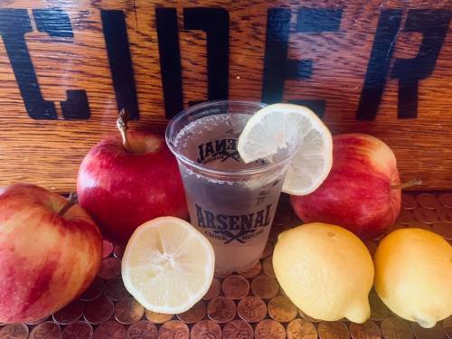 A clear cup of cider with lemon slices, surrounded by red apples and yellow lemons, against a wooden background.