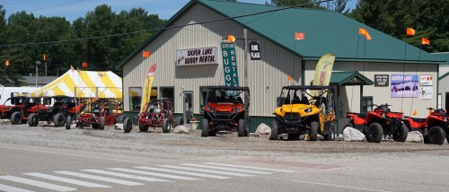 A row of ATVs and off-road vehicles parked outside a rental shop with a green roof and colorful flags.