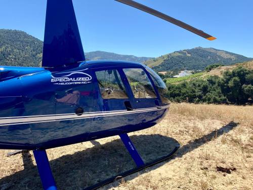 A blue helicopter parked on dry grass with mountains in the background under a clear blue sky.
