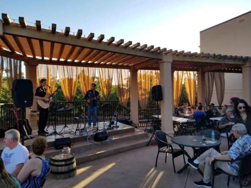 Two musicians perform on a stage under a pergola, while an audience enjoys the show at an outdoor venue.