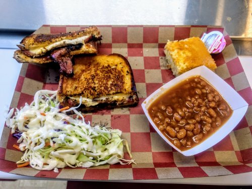 A tray with a grilled sandwich, baked beans, coleslaw, cornbread, and a small packet of sauce.
