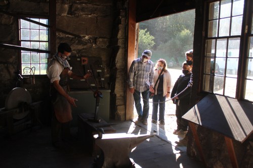 A blacksmith demonstrates his craft to a small group of onlookers in a sunlit workshop.