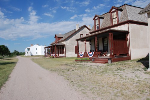 Historic buildings with porches line a dirt road under a blue sky, featuring decorative bunting.