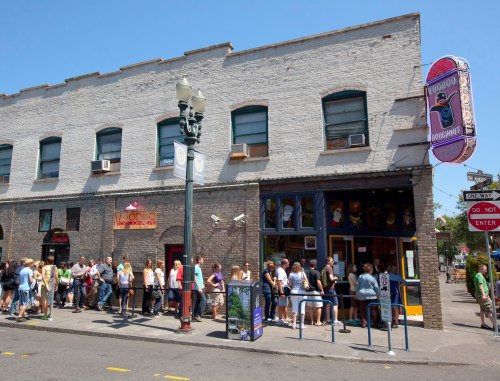 A long line of people waiting outside Voodoo Doughnut on a sunny day, with a colorful sign above the entrance.