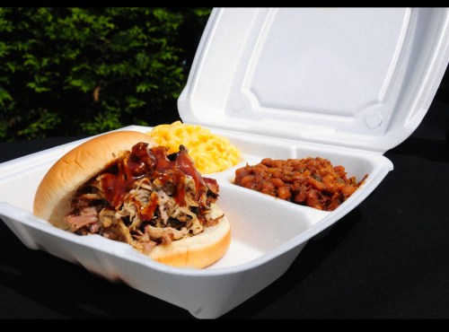 A takeout container with a pulled pork sandwich, macaroni and cheese, and baked beans.