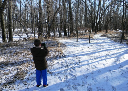 A child stands in a snowy forest, taking a photo of a trail sign that reads "No Exit Trail." Trees surround the area.