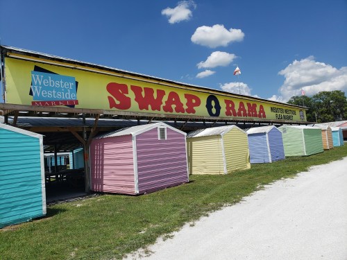 Colorful market stalls under a bright blue sky at Webster Westside Flea Market, with a large "SWAP O RAMA" sign.